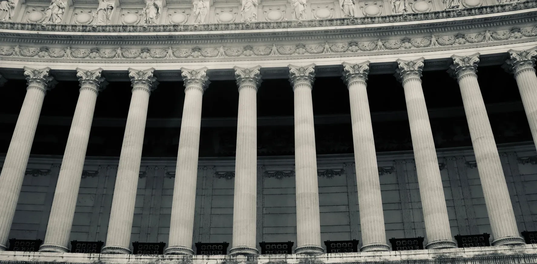 A row of tall neoclassical columns along the facade of a public institution, in muted black and white.